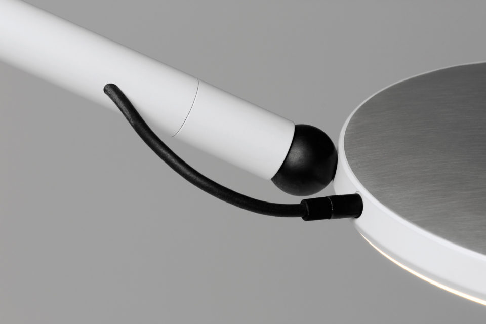 Quick-change Light Head System Section of an AVVENI luminaire, showing the tubular housing in white, from which a thin black cable protrudes and leads into the white luminaire head ring. The cover of the luminaire head is brushed and anodised silver.