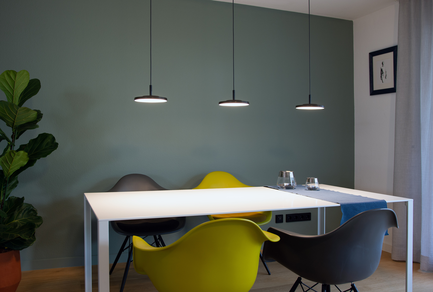 3 round, flat black lamp heads of the AVVENI Pendant 1 pendant luminaires above a white rectangular dining table with colourful chairs in front of a green wall.