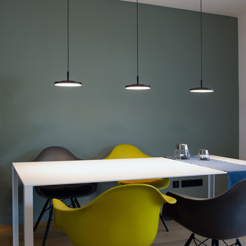 3 round, flat black lamp heads of the AVVENI Pendant 1 pendant luminaires above a white rectangular dining table with colourful chairs in front of a green wall.
