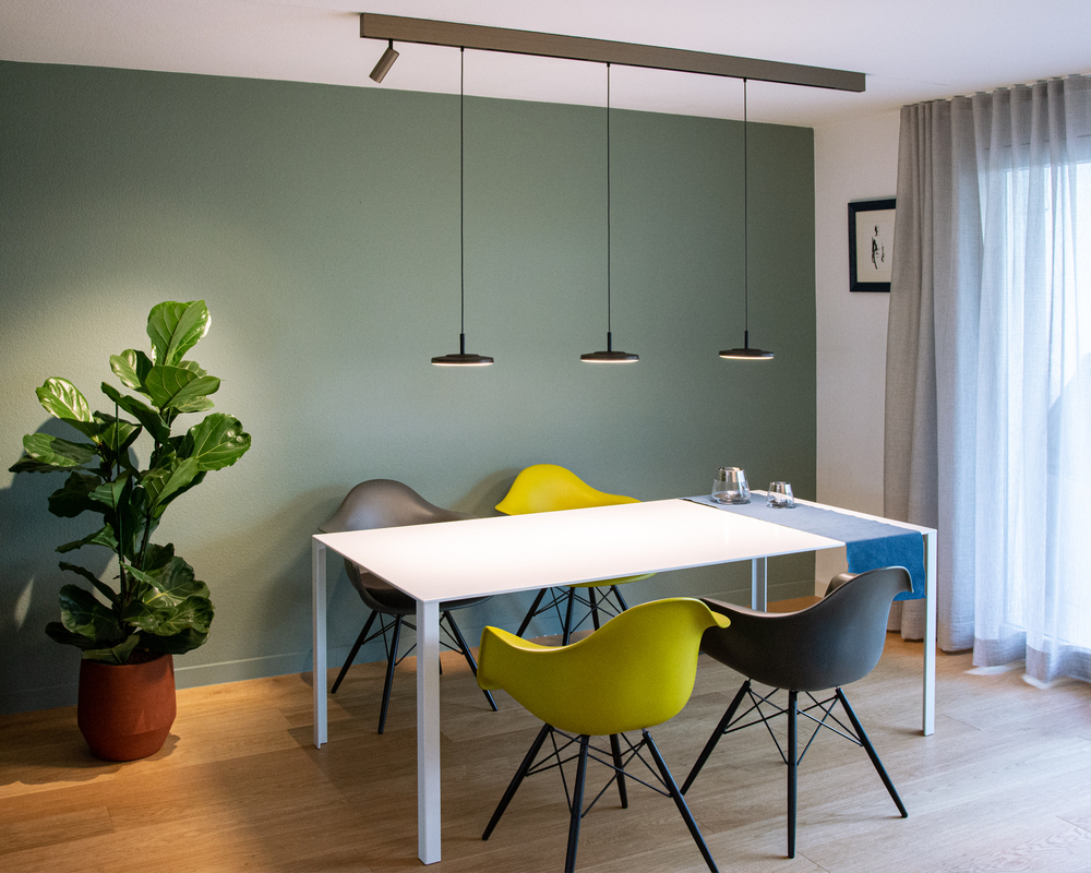 Lighting Element pendant luminaire AVVENI PENDANT 1 for PALADIO TRACK Modern dining room with white rectangular table and 4 colourful chairs around it. A track is attached to the ceiling above the table, into which a cylindrical spotlight (EVOLUTION SPOT S) is inserted on the left-hand edge, which illuminates the plant in the left-hand corner of the room, and 3 black pendant luminaires with round, flat lamp heads (AVVENI PENDANT 1), which illuminate the table.
