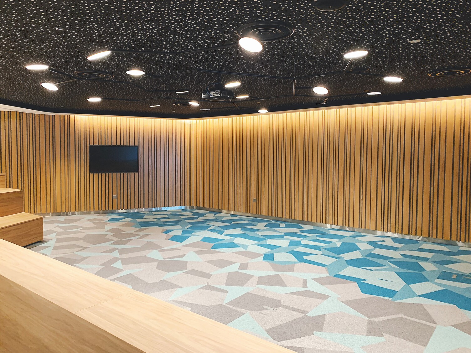 Reception desk in Capital Tower L9 Singapore with wooden counter, blue carpet and a black branched AVVENI Pendant 18 pendant luminaire with 18 round flat luminaire heads in front of a black ceiling.