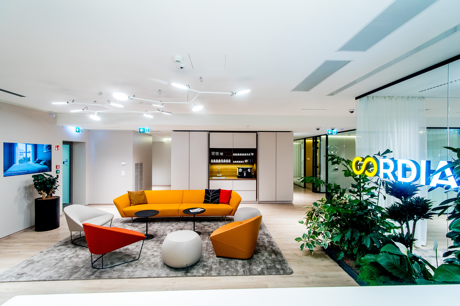 Waiting lounge with orange upholstered furniture above two white pendant luminaires, with a branched luminaire housing, the arms ending in flat round luminaire heads (AVVENI PENDANT), CORDIA logo on the right.