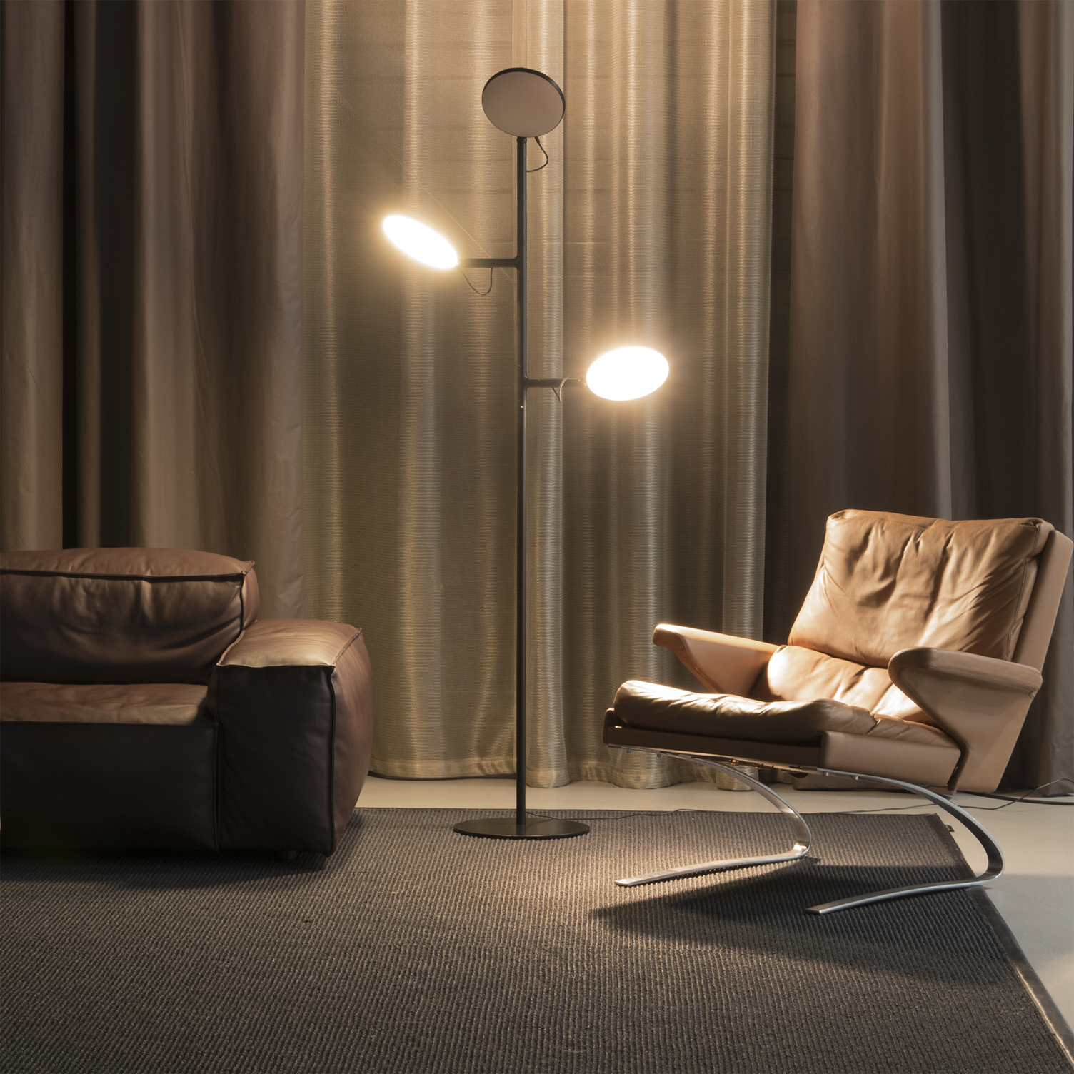 Between the brown leather sofa and leather armchair stands a black floor lamp with a round base from which a triple-branched cylindrical lamp base extends vertically upwards to the flat round lamp heads, the silver or gold lamp heads are partially turned from behind with the light side facing the black wall.