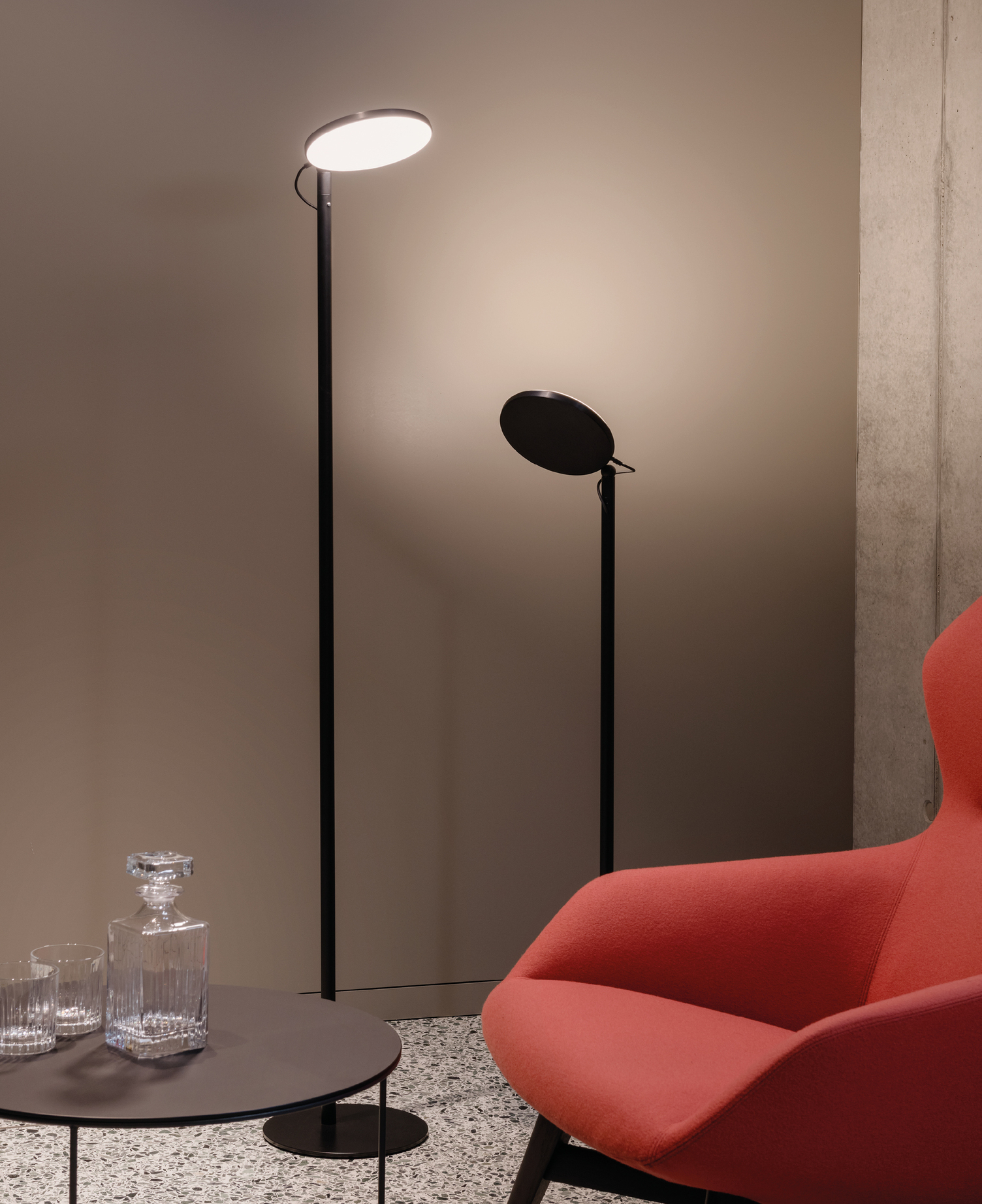 Two black floor lamps from the AVVENI FlOOR series of different heights with a round, flat lamp head behind a red armchair and table from the Brunner Group.