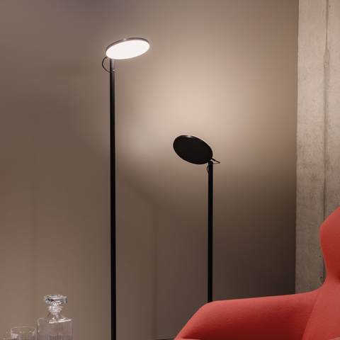 Two black floor lamps from the AVVENI FlOOR series of different heights with a round, flat lamp head behind a red armchair and table from the Brunner Group.