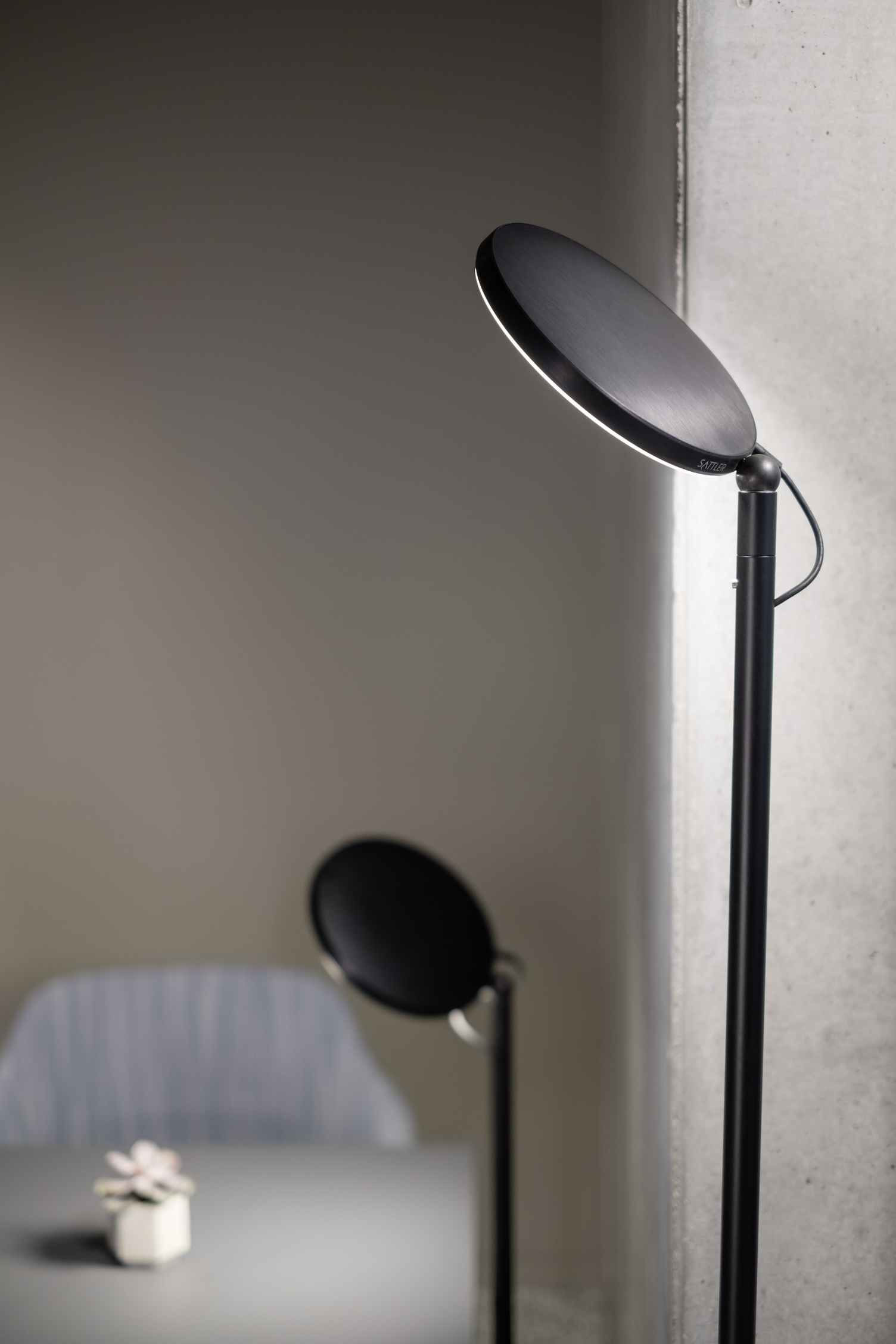 Two black floor lamps from the AVVENI FlOOR series of different heights with a round, flat lamp head at the side of a table and blue chair from the Brunner Group.