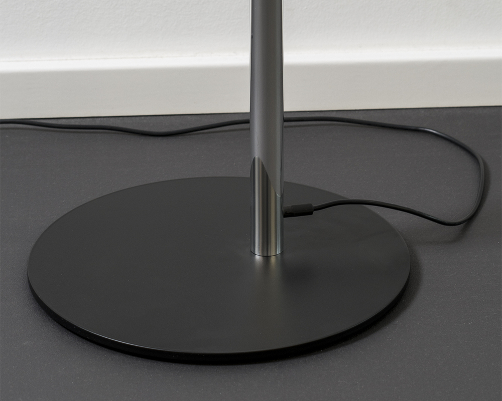 Flacher, runder Leuchtenfuß Auf schwarzem Boden, ein flacher runder schwarzer Metallfuß der Stehleuchte AVVENI FLOOR mit außermittigem zylindrischen Standfuß aus dem ein schwarzes dünnes Kabel herausragt und entlang der weißen Wandkante aus dem Bild entlangläuft.