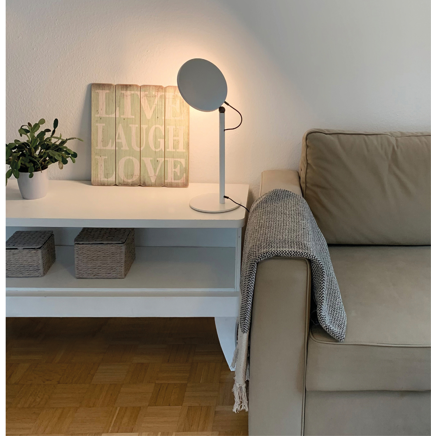 AVVENI TABLE table lamp as a decorative element in the living room Light for Live, Laugh, Love. White sideboard with 2 rectangular baskets, on which stands a plant and a green sign reading ‘Live, Laugh, Love’, illuminated by a white floor lamp, AVVENI TABLE, with movable round lamp head. Next to it is a beige sofa with a woollen blanket over the backrest.