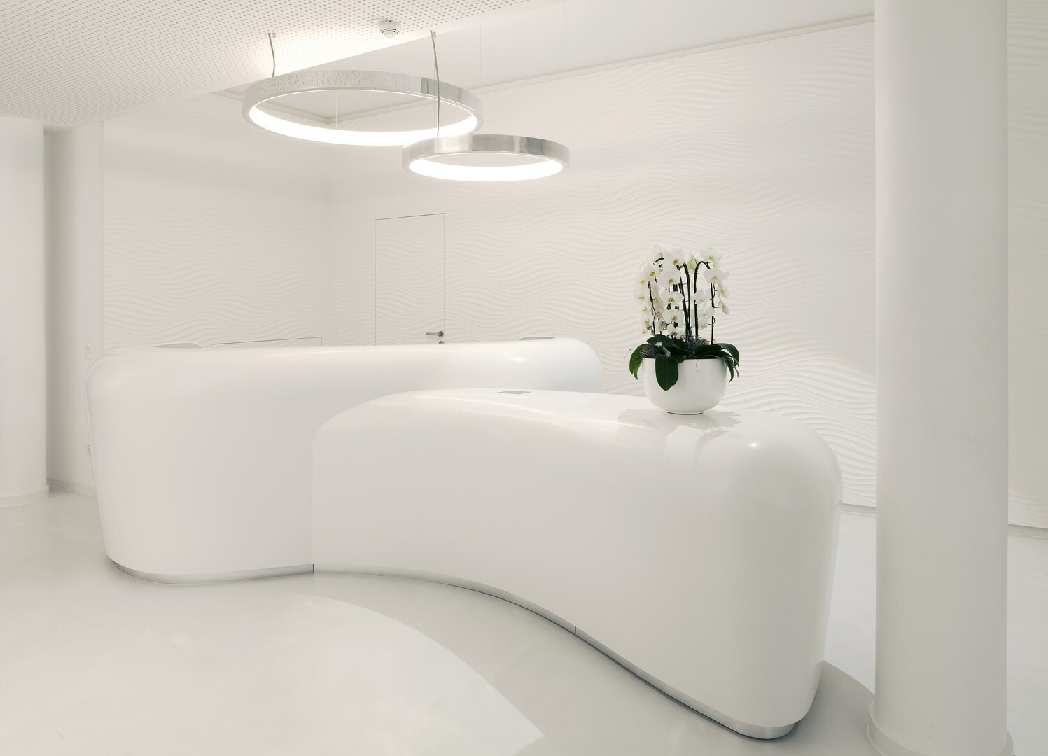 Ringleuchte für futuristische Empfangstheke Weißer Raum mit zwei weißen futuristischen Empfangstheken mit Orchideen auf einer, über den Theken sind 2 Aluminium Ringleuchten CIRCOLO abgependelt sind.