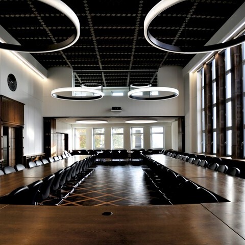 arge meeting room of the Duisburg Chamber of Industry and Commerce with dark furniture, wooden table arranged in a U-shape, with several large ring lights (CIRCOLO & CIRCOLO INVERSE.) with a diameter of 2.6 metres, suspended from the ceiling or surface-mounted.