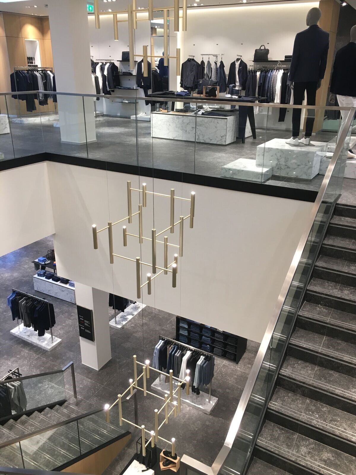 Gerade mehrläufige Steintreppe mit modernem Kronleuchter Blick auf eine Pendelleuchte bestehend aus drei modernen goldenen Kronleuchtern (EVOLUTION VERTICAL 3), in einem Steintreppenhauses mit Glaswangen eines Textilgeschäftes für moderne, edle Herrenbekleidung.