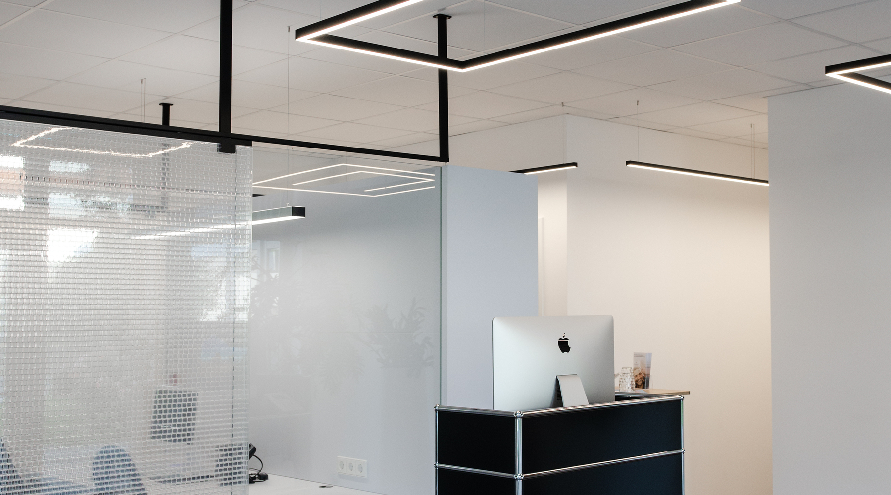 Kleiner rechteckiger Empfangstresen mit viereckigen Pendelleuchten GIOCO Kleiner schwarzer Empfangstresen vor einem Büro, das mit einer Glaswand abgetrennt ist, weiße Wände und weiße Decke, von der viereckige Pendelleuchten der GIOCO Serie abgehangen sind, zwei leuchten direkt weiß und eine indirekt in RGB Blau gegen die Decke.