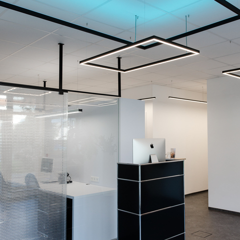 Small rectangular reception counter with square pendant lights GIOCO Small black reception desk in front of an office separated by a glass wall, white walls and white ceiling from which square pendant luminaires from the GIOCO series are suspended, two shine directly in white and one indirectly in RGB blue against the ceiling.