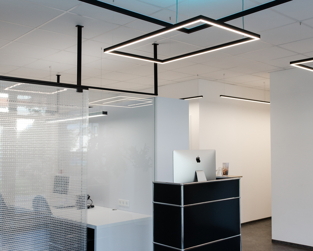 Kleiner rechteckiger Empfangstresen mit viereckigen Pendelleuchten GIOCO Kleiner schwarzer Empfangstresen vor einem Büro, das mit einer Glaswand abgetrennt ist, weiße Wände und weiße Decke, von der viereckige Pendelleuchten der GIOCO Serie abgehangen sind, zwei leuchten direkt weiß und eine indirekt in RGB Blau gegen die Decke.