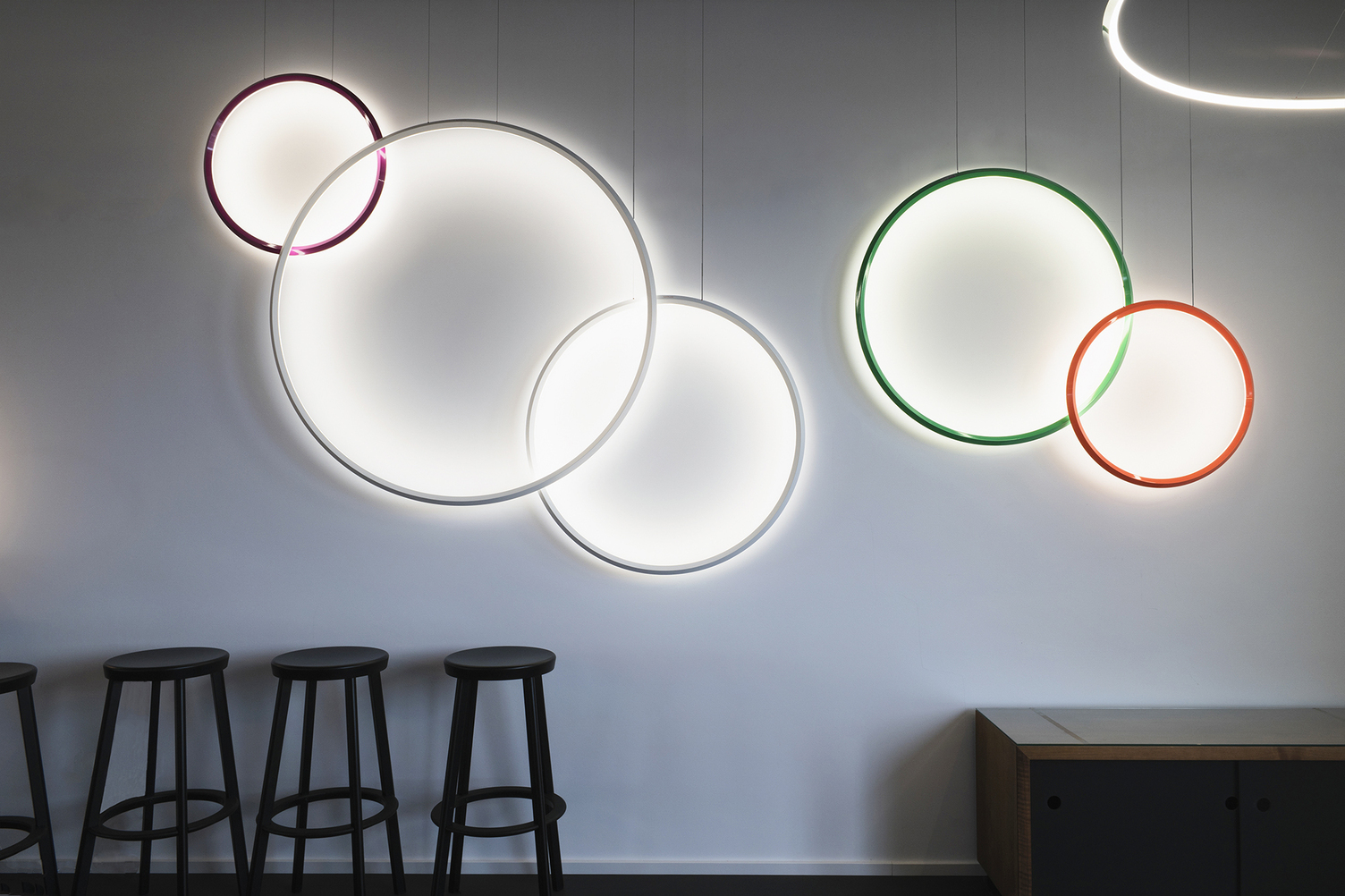 Surface colour variety Five ring lights from the GIRATA VERTICAL series in various bright colours – purple, white, red and green – and in various ring diameters are suspended at different heights along a white wall, shining into the centre of the circle and illuminating the wall at the same time. Several black bar stools are lined up to the left below them.