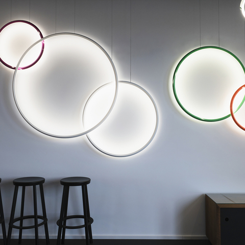 Surface colour variety Five ring lights from the GIRATA VERTICAL series in various bright colours – purple, white, red and green – and in various ring diameters are suspended at different heights along a white wall, shining into the centre of the circle and illuminating the wall at the same time. Several black bar stools are lined up to the left below them.