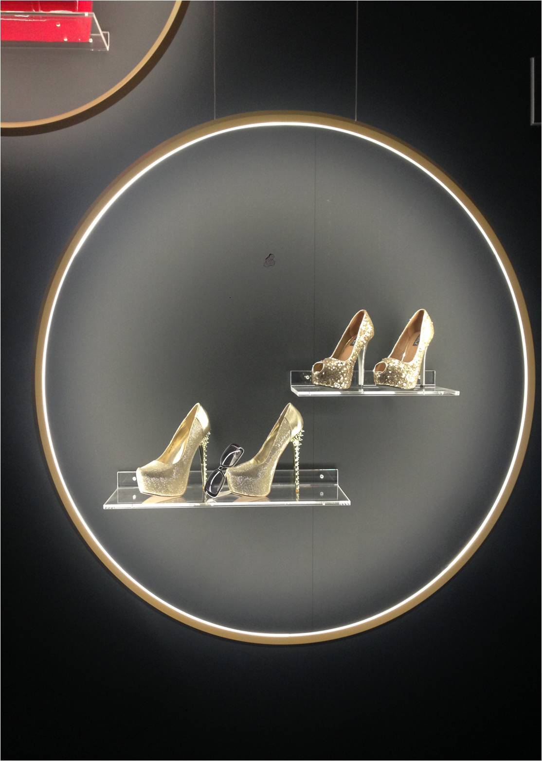 Ringleuchte GIRATA VERTICAL setzt Schuhe in Szene Innenkreis der Ringleuchte GIRATA als Präsentationsfläche für goldene Schuhe & schwarze Sonnenbrille auf Regalen vor einer schwarzen Wand.