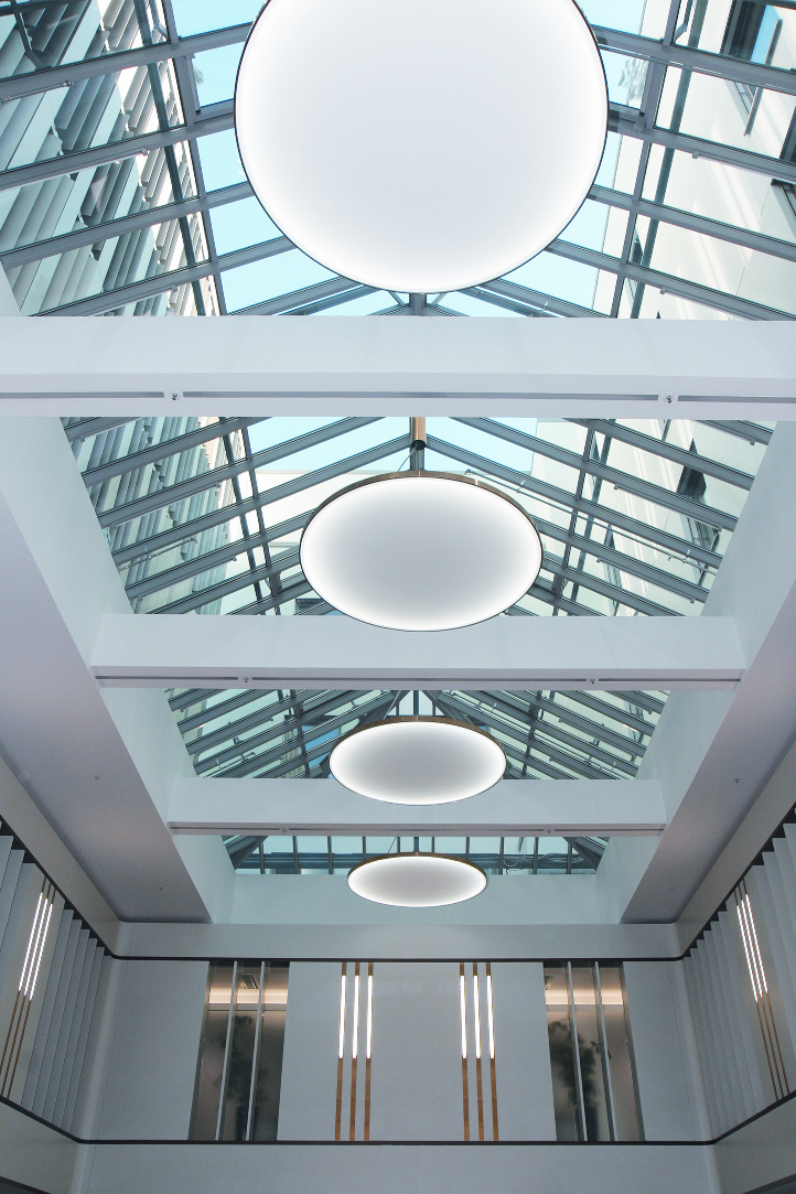 Blick von unten auf runde flächige Leuchten, abgependelt in einem Atrium mit spitzem Glasdach.
