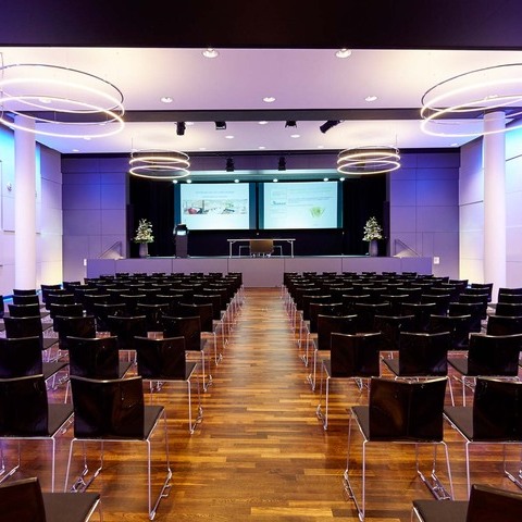 Bananacity event hall with 4 INSIEME chandeliers Bananacity event room in Switzerland with a view of the stage, block seating in front of it. Room with white walls coloured purple by light, an INSIEME ring chandelier hangs in all corners of the room.