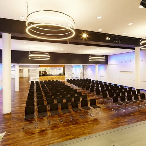 Bananacity event hall with 4 INSIEME chandeliers Bananacity event room in Switzerland with a view from the stage onto the hall with block seating and round white pillars. The room has white walls coloured purple by light and an INSIEME ring chandelier hangs in all 4 corners of the room.