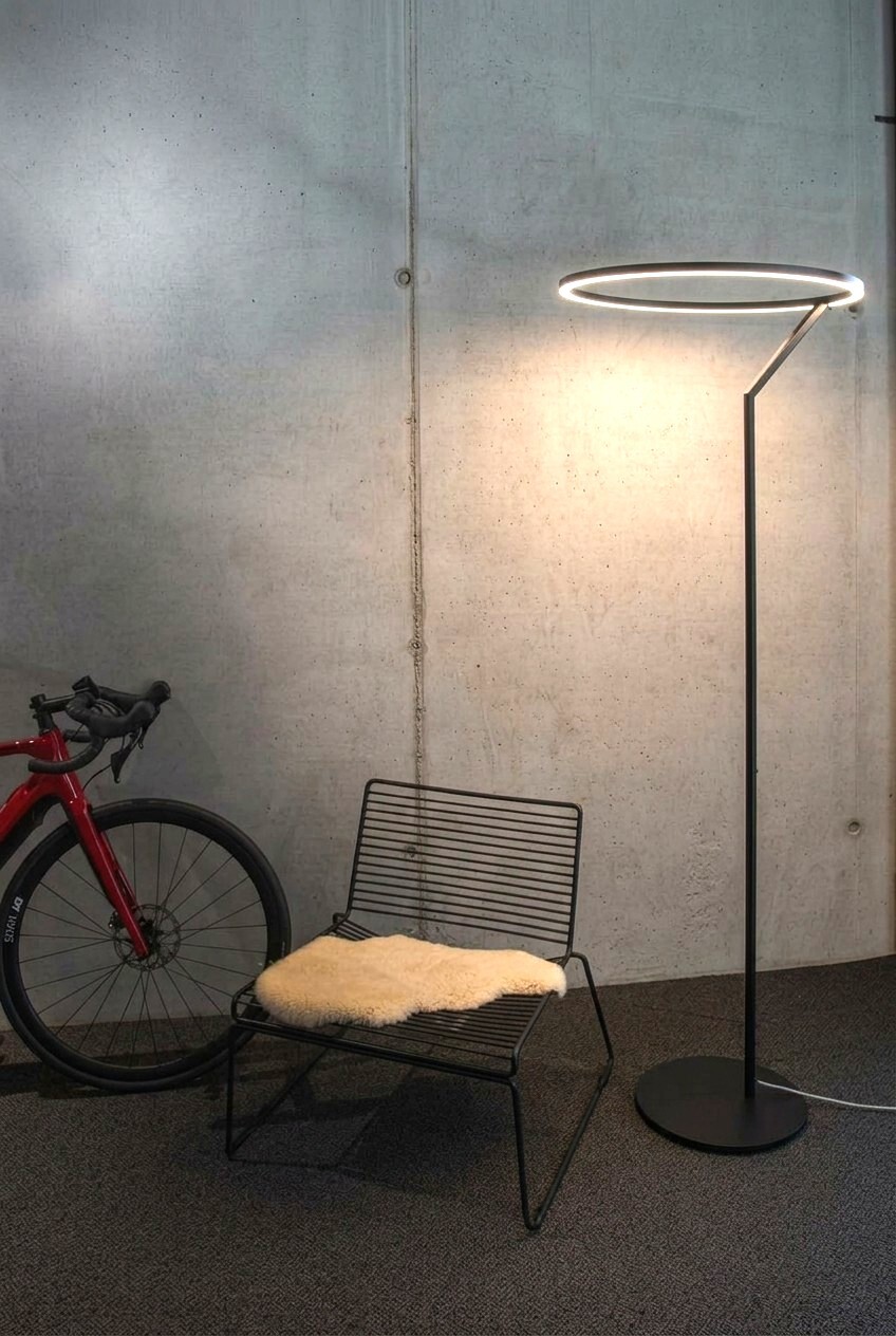 INSOSPESO FLOOR - Elegante Metall-Stehleuchte mit Ringlicht & besonderer Formenführung Betonwand an der von links ein rotes Fahrrad steht, daneben ein moderner Metallstuhl, daneben steht eine große Stehleuchte, die Insospeso Floor: Gerader schmaler Metallfuß mündet oben in einen leuchtenden Ring, der nach unten strahlt.