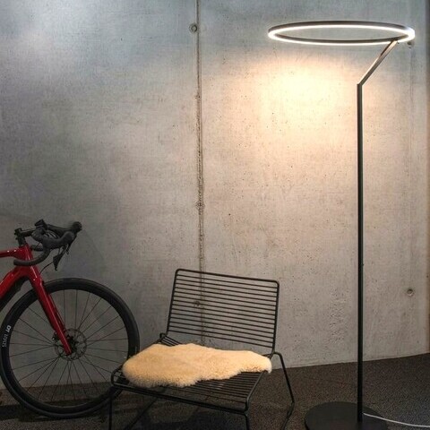 INSOSPESO FLOOR - Elegante Metall-Stehleuchte mit Ringlicht & besonderer Formenführung Betonwand an der von links ein rotes Fahrrad steht, daneben ein moderner Metallstuhl, daneben steht eine große Stehleuchte, die Insospeso Floor: Gerader schmaler Metallfuß mündet oben in einen leuchtenden Ring, der nach unten strahlt.