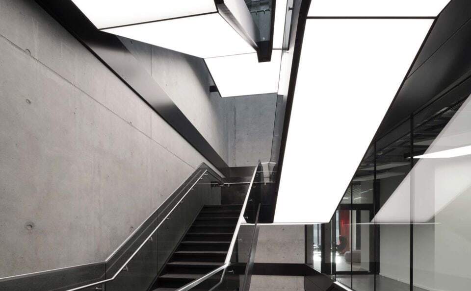 Modern staircase in an exposed concrete building, integrated luminous ceilings run along the underside of the staircase in square, divided strips and in turn illuminate the steps and corridors below.