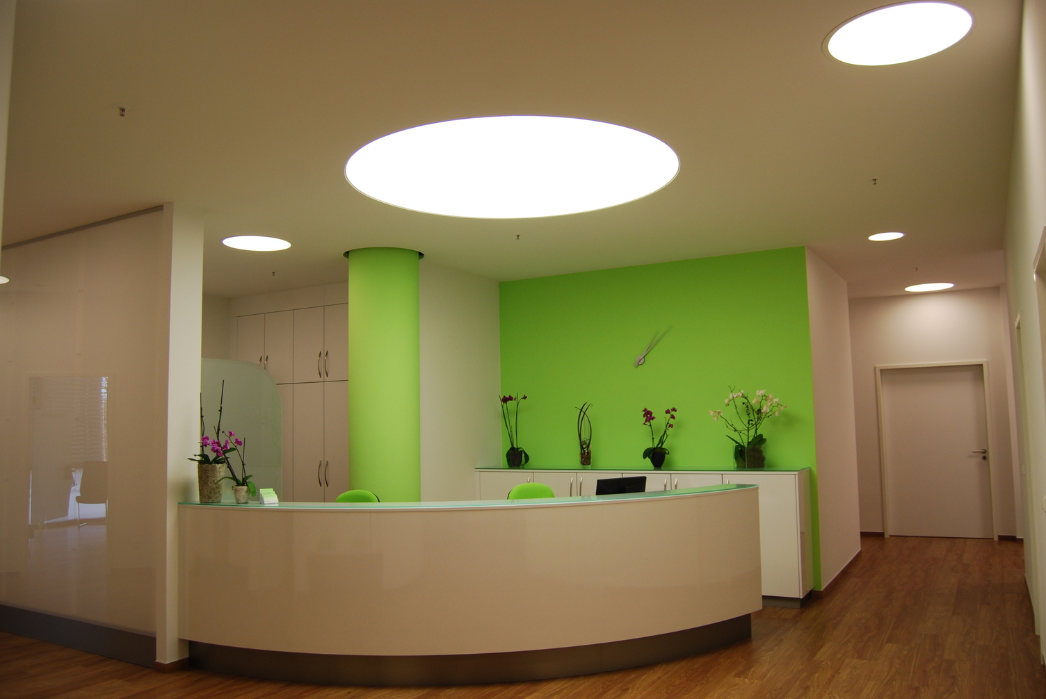 Runde Einbaulichtdecke über runder Empfangstheke Runde weiße Hochglanz-Empfangstheke, die an einer Wand anschließt. Die Wand und die Stühle sind in grellem grün gehalten. Über der Theke ist eine große runde deckenschlüssige Einbaulichtdecke von SATTLER und beleuchtet die Theke und die dahinter liegenden Arbeitsplätze.