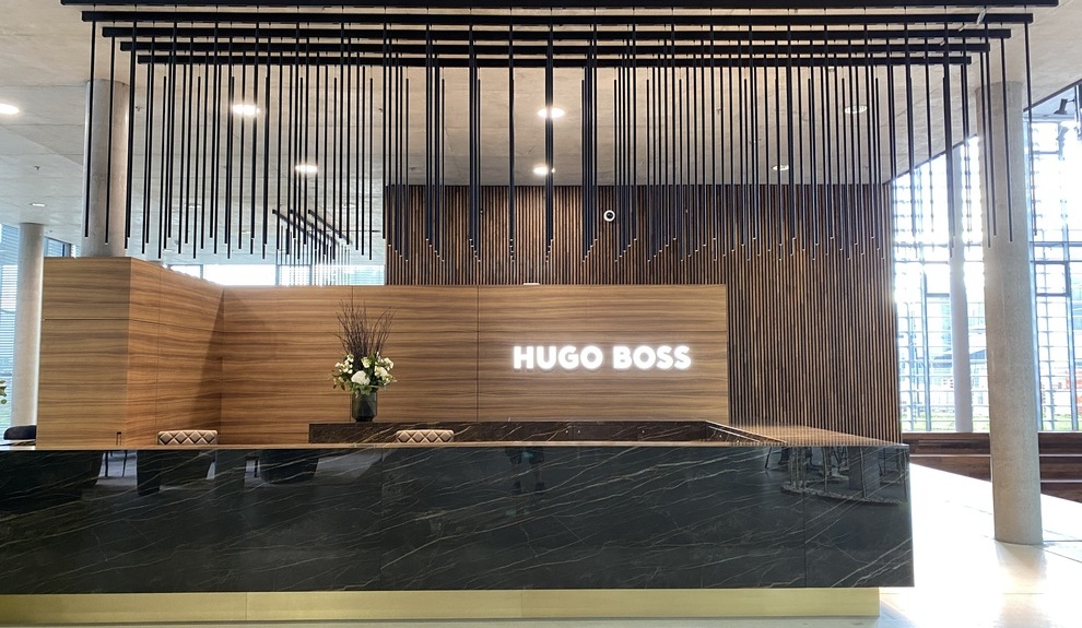 Zylindrische Pendelleuchten FINO schmücken Empfangstresen Dunkler eckiger edler Marmorempfangstresen mit Holzrückwand auf der leuchtend weiß das Hugo Boss Logo steht. Über dem Empfangsbereich sind parallel mehrere PALADIO TRACK Stromschienen an der Decke angebracht, von denen lange zylindrische Leuzchten abgependelt sind.