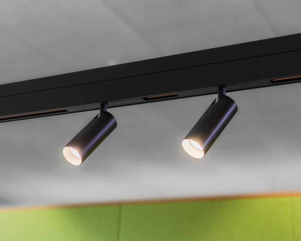 Movable cylindrical EVOLUTION SPOT S as a lighting element for PALADIO TRACK Section of a black linear track luminaire PALADIO TRACK with two inserted, movable, black, cylindrical spots (EVOLUTION SPOT S), which shine obliquely forwards, in front of a green felt wall.