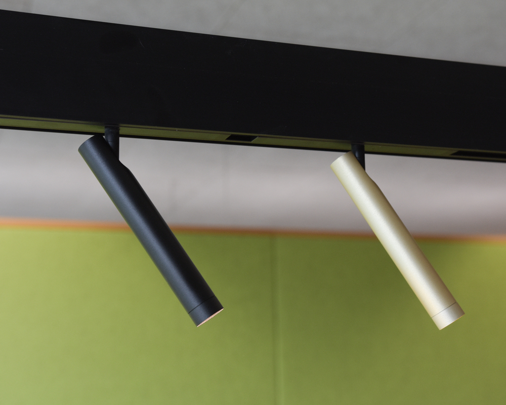 Movable slim cylindrical FINO SPOT 2 as a lighting element for PALADIO TRACK In front of a green felt wall: section of a black linear track luminaire PALADIO TRACK with two inserted movable, slim cylindrical spots (FINO SPOT 2) in black and champagne gold, which shine obliquely forwards to the right.