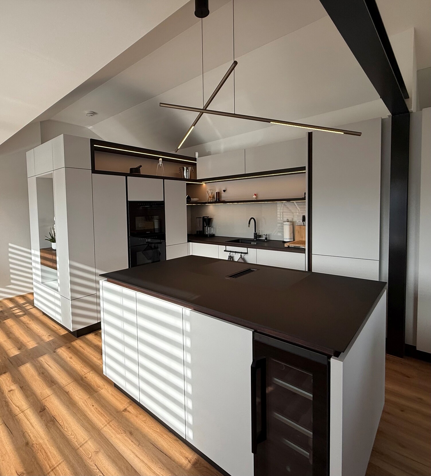 PALITO als Pendelleuchten Küchenblock Küchenlampen. Moderne private offene Küche mit Holzboden, das moderne Mobiliar, Küchenschränke und der Küchenblock in der Mitte sind in weiß und mit schwarzen Elementen. Über dem freistehenden Küchenblock sind zwei zylindrische, waagerechte PALITO Pendelleuchten.