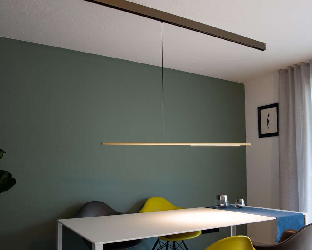 PALITO for PALADIO TRACK (track system) Dining room with a white rectangular table surrounded by yellow and grey shell chairs, set against a green wall. A cone of light can be seen on the table, emitted by a cylindrical, horizontally suspended luminaire, which in turn is suspended from the ceiling by a surface-mounted track.