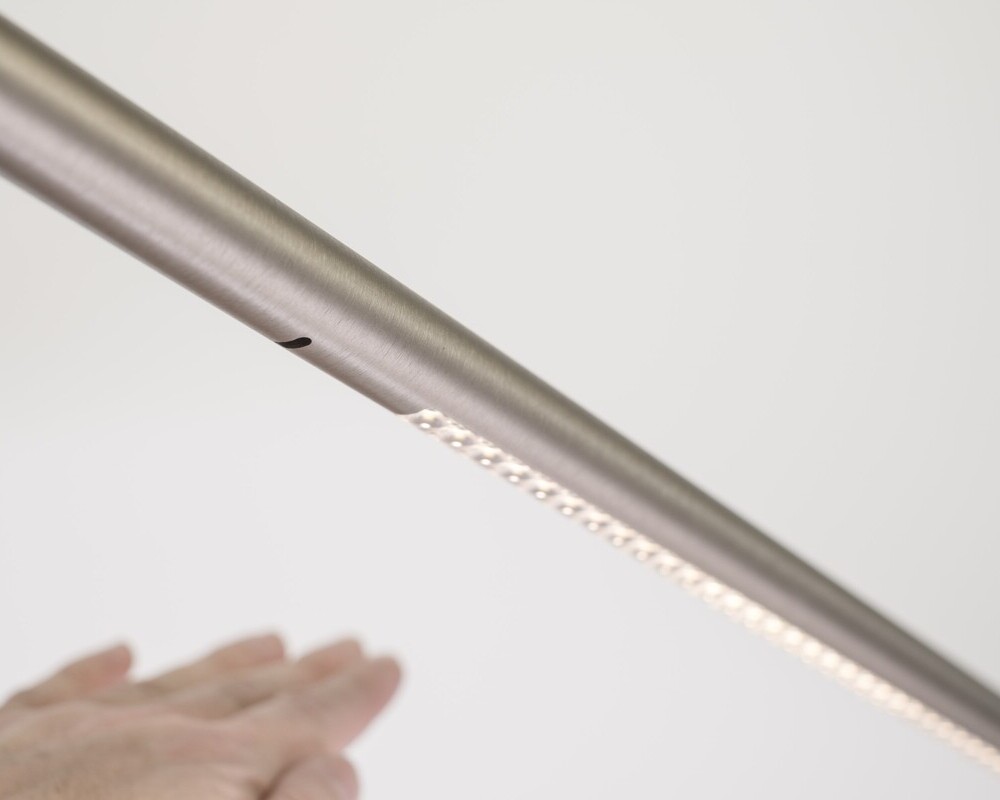 Gesture control Cylindrical aluminium luminaire running diagonally horizontally, a hand can be seen under a small hole in the tube, illuminated lenses can be seen in the further course of the cylindrical luminaire on the right. The picture shows the gesture control and dimming of the luminaire.