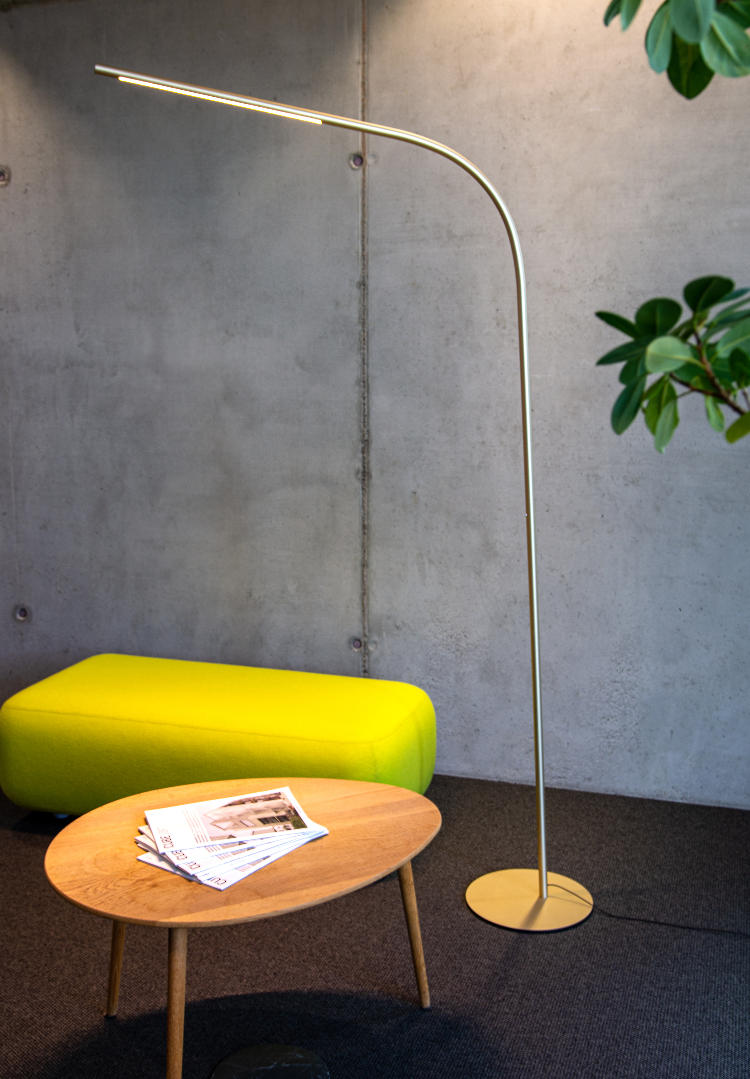 Gebogene Lounge-Stehleuchte Moderne Betonwand vor der eine knall-gelbes strahlende Sofasitzbank steht. Davor ein ovaler Holztisch. Neben einer großen grünen Zimmerpflanze ragt eine zylindrische gebogene hohe Lounge-Stehleuchte heraus. Die PALITO FLOOR bringt ein edles Ambiente mit nicht zuletzt durch die hochwertige Oberflächenfarbe in eloxiertem Champagner-Gold.