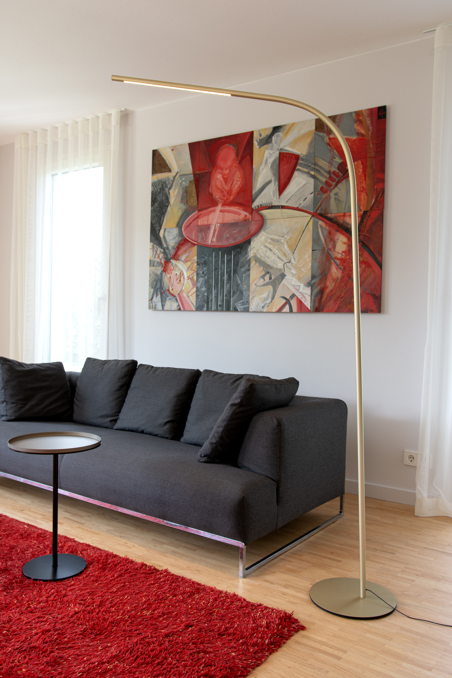 Wohnzimmerstehleuchte PALITO FLOOR Wohnzimmer mit rotem Teppich und großem Bild an der Wand in Rottönen. Eine große dunkelgraue Sofalandschraft erstreckt sich entlang der Wand. Am Rand des Sofas steht eine gebogene große schmale Stehleuchte PALITO FLOOR in schönem Champagner-Gold für das perfekte, dimmbare Leselicht.