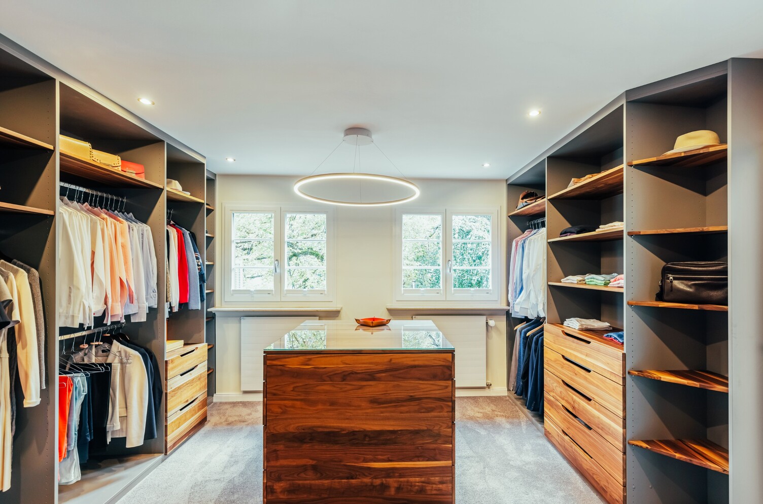 Ankleidezimmer mit Ringpendelleuchte SLIM DIRECT Moderner Ankleideraum mit links und rechts vom Raum Regalen und Schränken mit Kleidung in der Mitte ein Holz-Tischblock über dem eine Ringleuchte SLIM DIRECT hängt.