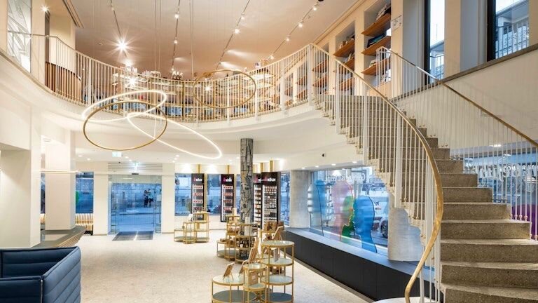 Köln Tourismus mit einem Kronleuchter aus SLIM Ringleuchten | © Köln Tourismus Blick vom Innenraum der Köln Tourismus auf eine Treppe, die in den ersten Stock führt und den Glaseingang, in der Mitte des Raumes zieht ein Lichtobjekt aus SLIM-Ringleuchten die Blicke auf sich. | © Köln Tourismus