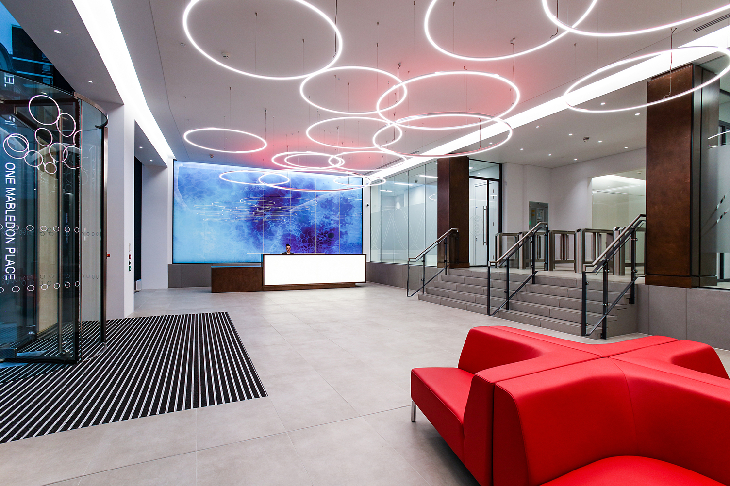 Company entrance area with several SLIM ring lights The entrance area of One Mabledon Place in London shows a tiled entrance area above which several SLIM ring lights are suspended. At one end of the room is a reception desk behind which hangs a large blue picture, a red leather sofa on the other side of the room, stairs lead up one level from the glass entrance door.