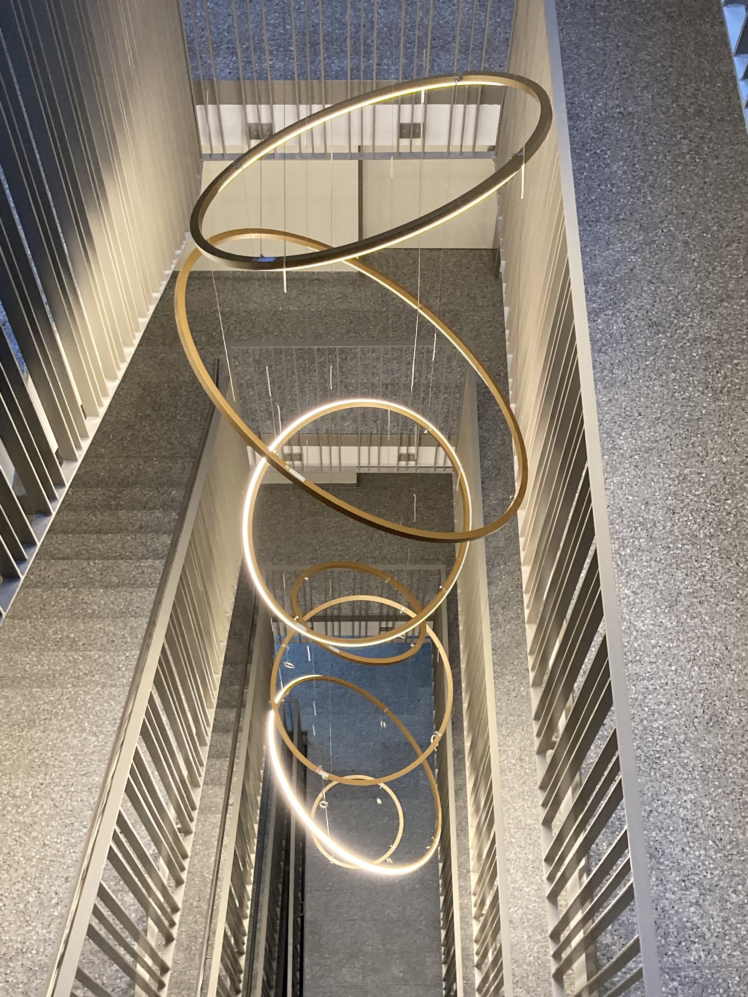 Beleuchtung Treppenauge - Kronleuchter aus 7 SLIM Ringleuchten Blick nach unten auf eine Sonderleuchte, wie ein moderner Kronleuchter bestehend aus mehreren direkt leuchtenden Ringleuchten, in einem Treppenauge eines Treppenhauses mit mehrläufigen geraden Treppen.