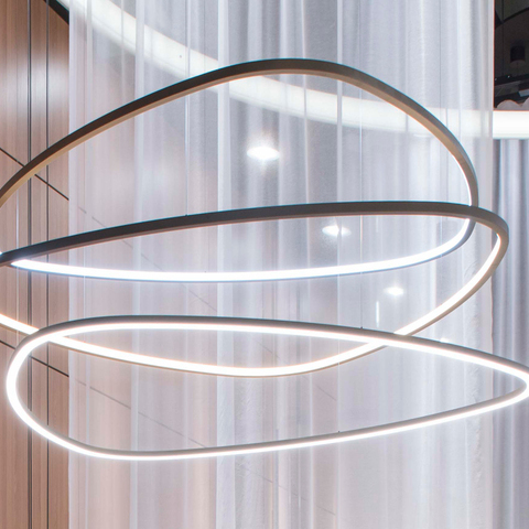 SLIM DELTA triangular ring lights with direct or inward illumination In the foreground, three narrow ring lights in the shape of a rounded triangle are suspended a short distance above each other, with the lowest one shining directly downwards and the two upper ones shining into the centre of the lights. In the background, a brown wooden wall and a slightly transparent white curtain, and a modern TOCCATA ring chandelier.