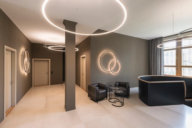 Waiting area with SLIM ring lights Small waiting area with sofa furniture in an earth-coloured hallway with an angular column, to the right a spiral staircase, on the walls several SLIM WALL ring luminaires of different diameters and delicate SLIM ring luminaires suspended from the ceiling.
