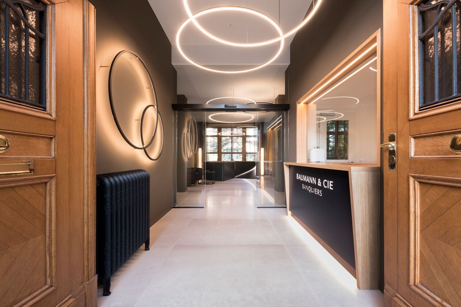 Eingangsflur einer Bank mit SLIM-Ringleuchten Historische zweiflügelige Holzeingangstür mit Ornamenten gibt Blick frei auf einen Flur in Holz und Erdtönen mit dekorativ angeordneten Slim Wall gegenüber eines Empfangs der Bank Baumann Cie und für die Grundbeleuchtung eingesetzte Slim direkt, sowie Slim indirekt leuchtenden Ringleuchten.