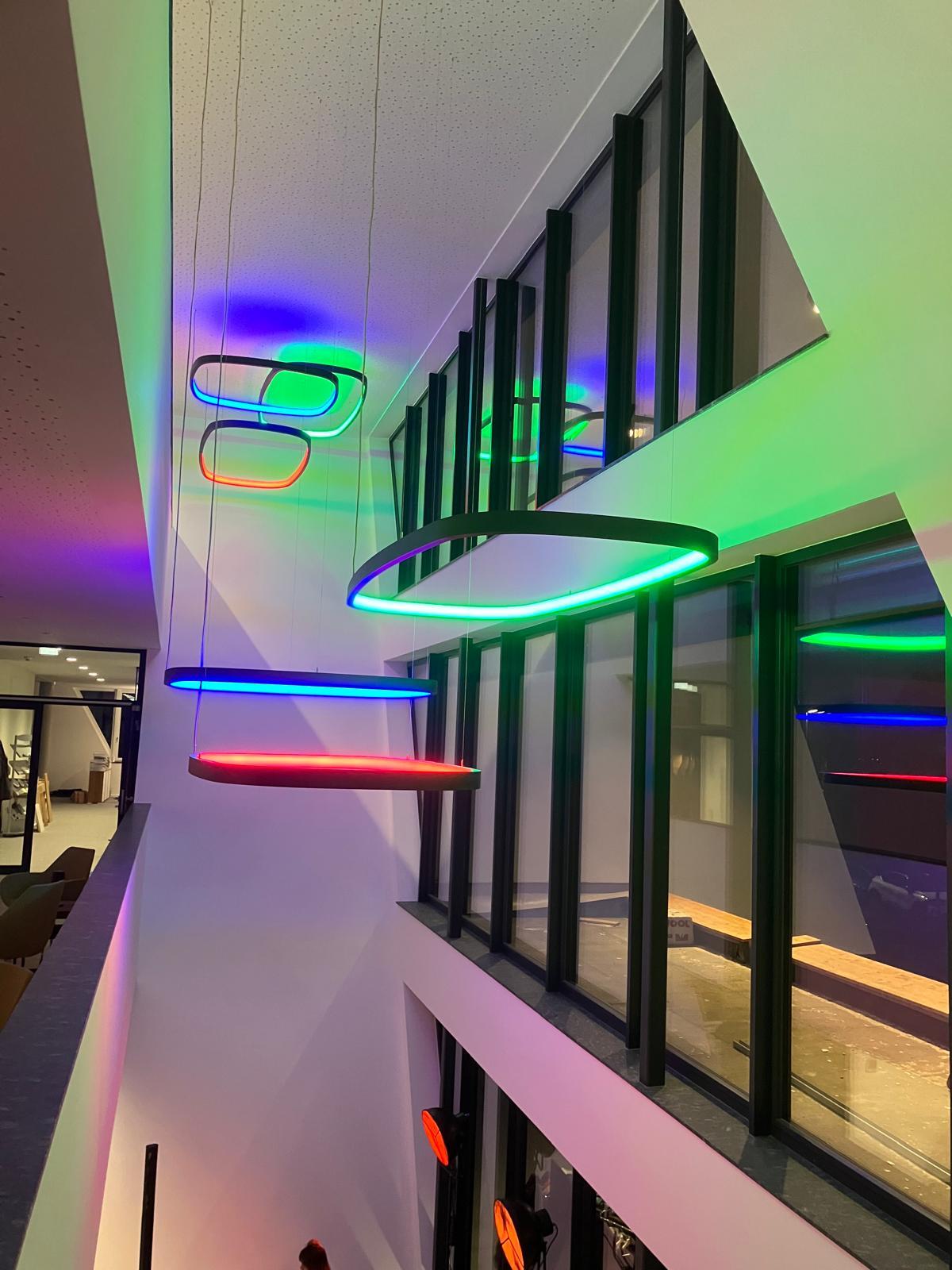 Großdimensionale Leuchten mit RGB in Treppenhaus / Atrium Innenraum eines modernen Gebäudes, links zeigt es einen Gang in höherem Stock, im Atrium/Treppenhaus, dass sich zu der Glasaußenfassade ergibt sind 6 großformatige viereckige, SOFT QUADRA, und rechteckige, SOFT SQUARE, Rahmenleuchten mit RGB in unterschiedlichen Höhen abgehangen, jede erstrahlt in unterschiedlicher Farbe: rot, grün, blau.