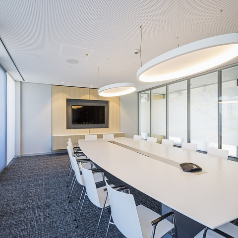Bright meeting room with elliptical acoustic pendant light SOFT ELLIPSE L+A A light-coloured meeting room with a rectangular white table and white chairs in front of a TV screen. Two large elliptical white acoustic lights, SOFT ELLIPSE L+A, are suspended above the table.