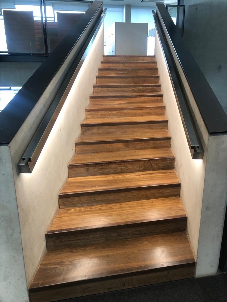Treppenstufenbeleuchtung unter Handlauf Gerade Treppe mit Trittstufen aus Holz, Seitenwände aus Beton, auf der Innenseite der Wände ist auf beiden Seiten ein Handlauf aus Metall angebracht, aus dem nach unten Licht auf die Stufen leuchtet.