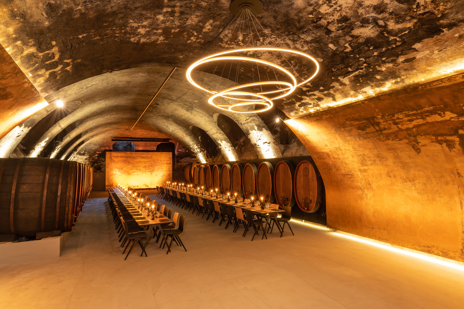 Chandelier TOCCATA in the wine vault cellar of the Staatliche Hofkellerei Würzburg Vaulted wine cellar of the Staatliche Hofkellerei Würzburg with wooden barrels along the walls, in the centre is a long, festive table consisting of several wooden tables and chairs with candles on the tables, above which hovers a modern chandelier consisting of several interconnected ring lights (TOCCATA).