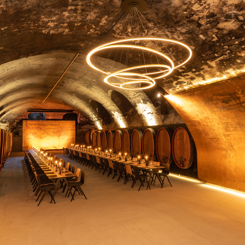 Chandelier TOCCATA in the wine vault cellar of the Staatliche Hofkellerei Würzburg Vaulted wine cellar of the Staatliche Hofkellerei Würzburg with wooden barrels along the walls, in the centre is a long, festive table consisting of several wooden tables and chairs with candles on the tables, above which hovers a modern chandelier consisting of several interconnected ring lights (TOCCATA).