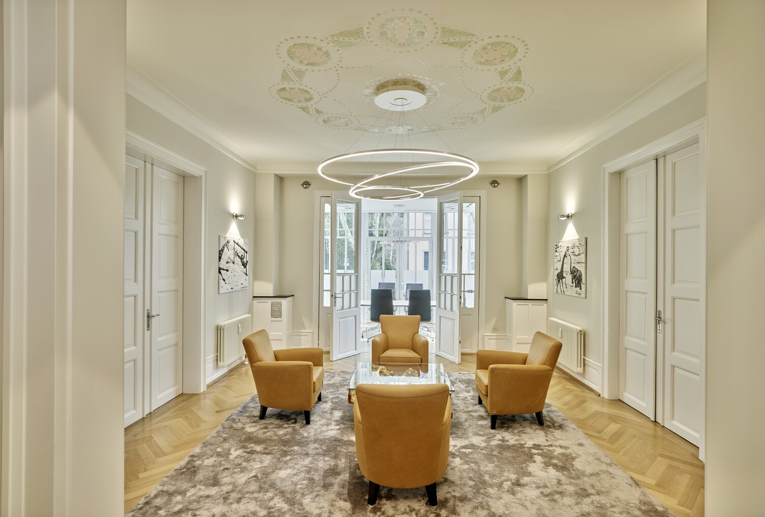 Wartebereich in einer klassischen Bürovilla mit 4 gelben Sesseln um einen Glastisch, über dem Glastisch schwebt ein moderner Ringleuchten-Kronleuchter (TOCCATA), der von einer verzierten Decke abgehangen ist.