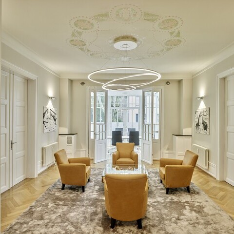 Waiting area in a classic office villa with 4 yellow armchairs around a glass table, above the glass table hovers a modern ring chandelier (TOCCATA) suspended from an ornate ceiling.